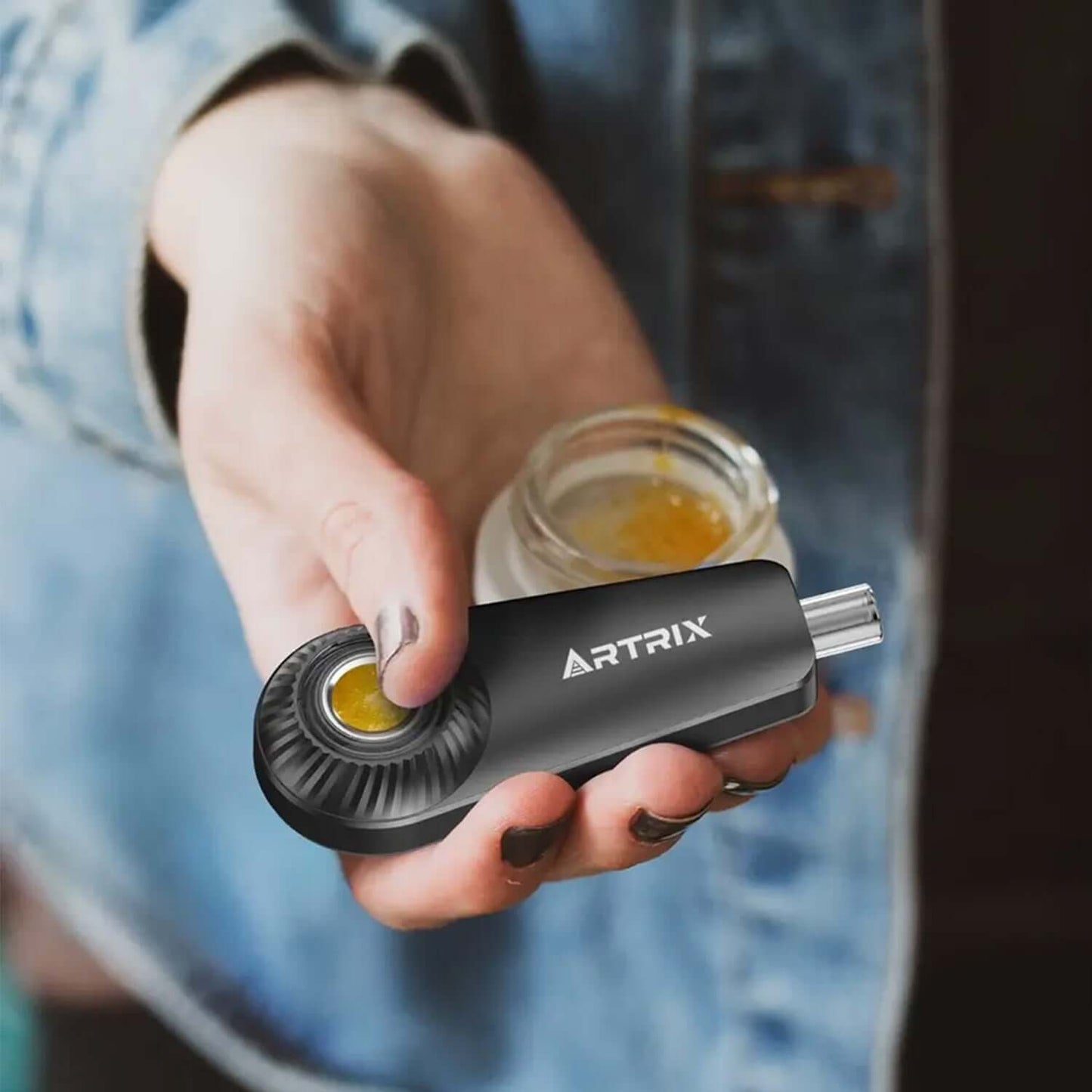 Hand holding ARTRIX portable dab pen with concentrate jar in the background, showcasing modern vaping experience.